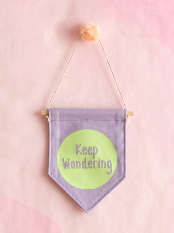 A purple canvas banner that reads 'Keep Wondering' in a green circle is hung on a pink wall.