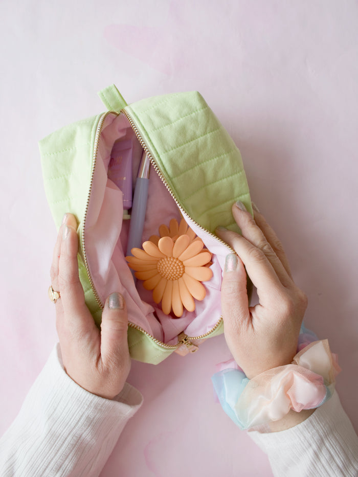 A pair of female hands holding open a green quilted zip pouch on a pink-marbled floor to reveal its content of hair clips, a brush and cream.
