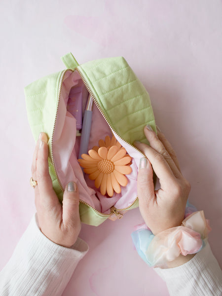 A pair of female hands holding open a green quilted zip pouch on a pink-marbled floor to reveal its content of hair clips, a brush and cream.