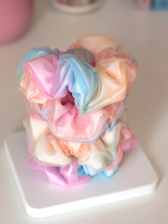 Four rainbow-coloured scrunchies are placed on top of each other on a white and clear holder.