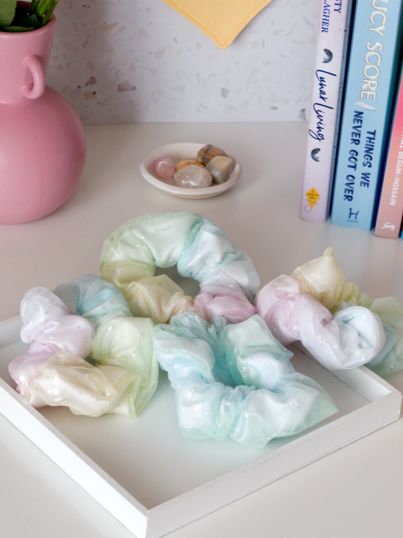 Four rainbow-coloured scrunchies are arranged on a white tray on a dressing table. Surrounding them are a vase, books and crystals.