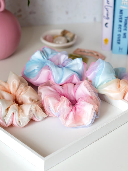 Four rainbow-coloured scrunchies are arranged on a white tray on a dressing table. Surrounding them are a vase, books and crystals.