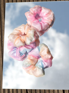 Four rainbow-coloured scrunchies are placed on a mirror where you can see the blue and cloudy reflection from the sky.