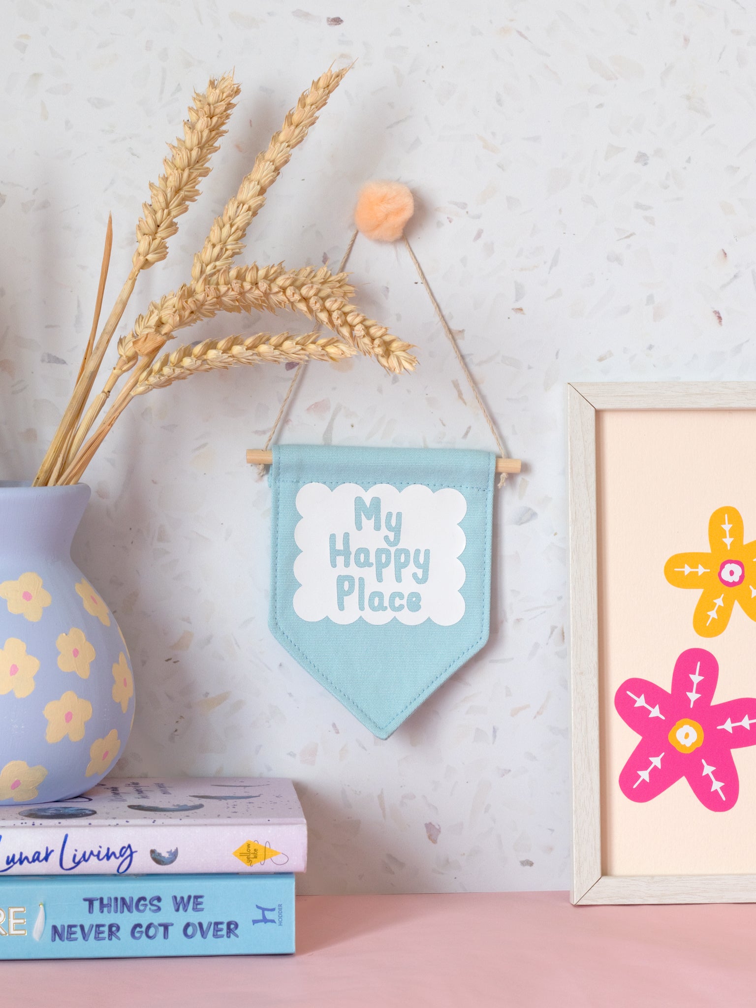 A blue canvas banner that reads My Happy Place in a white scalloped box is hung on a white terrazzo wall surrounded by framed art, flowers and books.