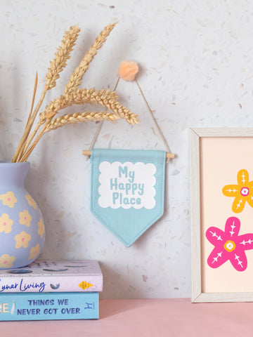 A blue canvas banner that reads My Happy Place in a white scalloped box is hung on a white terrazzo wall surrounded by framed art, flowers and books.