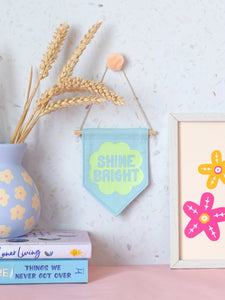 A blue canvas banner that reads Shine Bright in a green flower is hung on a white terrazzo wall surrounded by framed art, flowers and books.