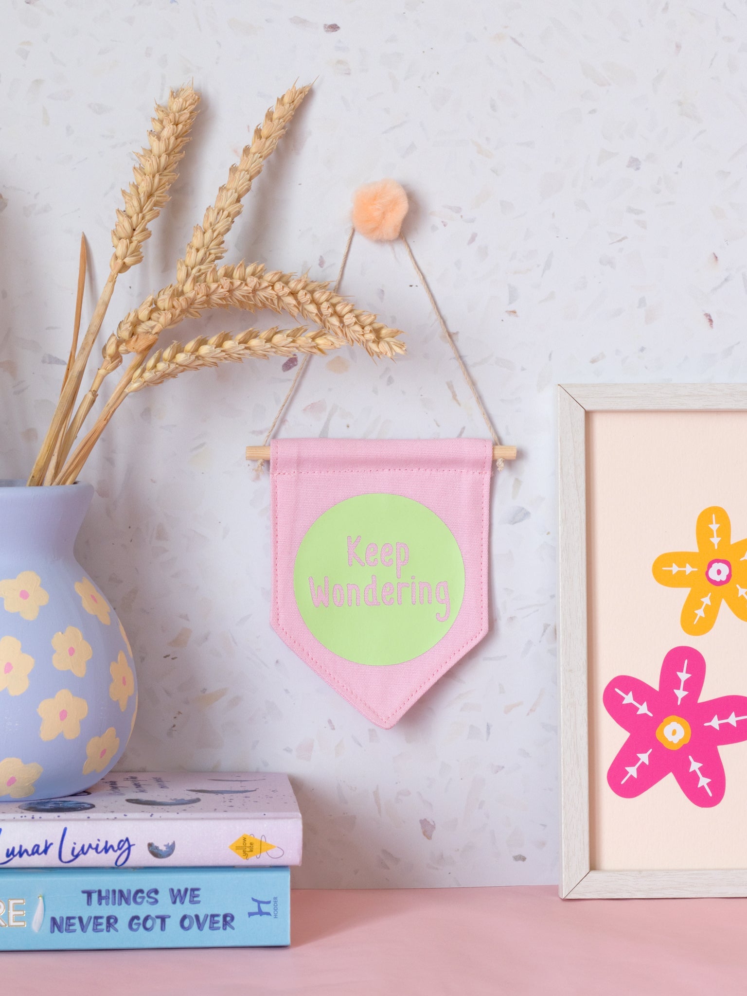A pink canvas banner that reads 'Keep Wondering' in a green circle is hung on a white terrazzo wall surrounded by framed art, flowers and books.