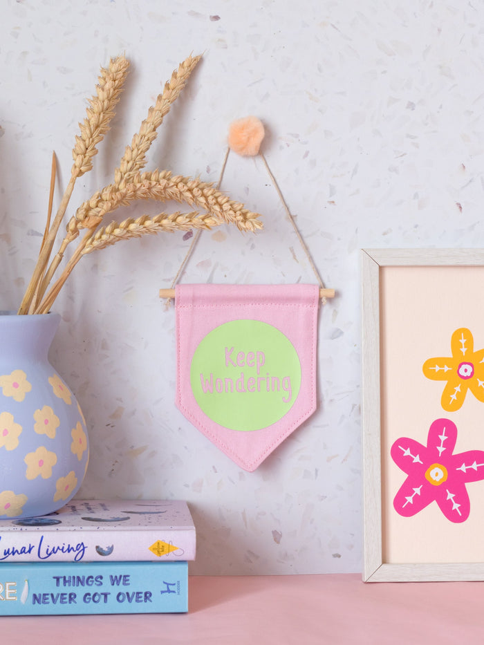 A pink canvas banner that reads 'Keep Wondering' in a green circle is hung on a white terrazzo wall surrounded by framed art, flowers and books.