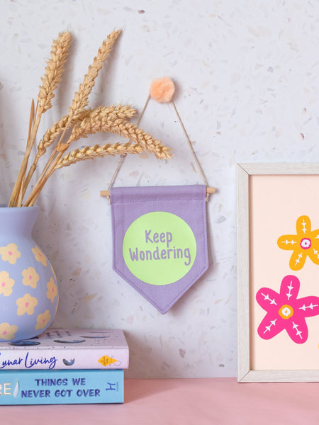 A purple canvas banner that reads 'Keep Wondering' in a green circle is hung on a white terrazzo wall surrounded by framed art, flowers and books.