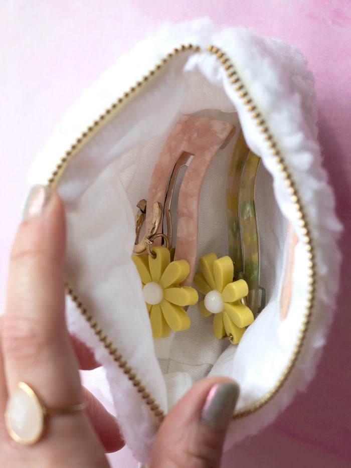 A white fluffy coin purse held open by female fingers to show its contents of resin hair clips and yellow flower earrings.