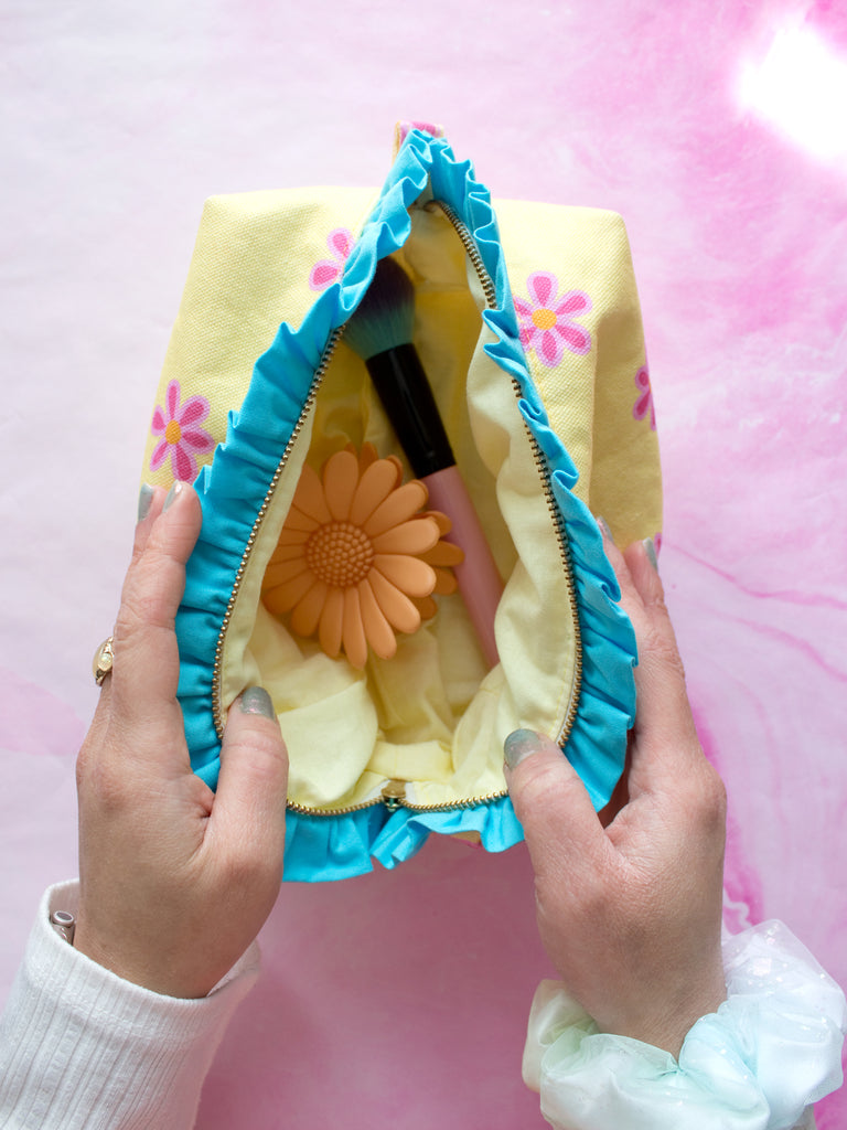 A pair of female hands holding open a yellow floral with blue ruffles zip pouch on a pink-marbled floor to reveal its content of hair clips and a brush.