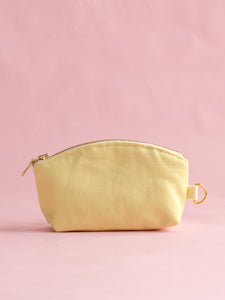 A yellow canvas coin purse with a metal zip and ring hook on a pink foreground.