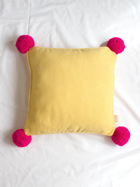 A yellow textured cushion with bright pink pom-poms in the centre of a white bedsheet.