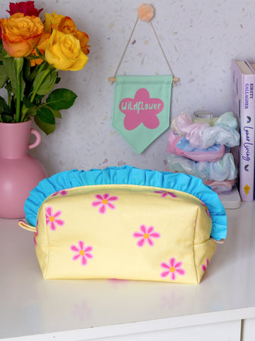 A yellow floral with blue ruffles zip pouch with a fluffy pink flower in the centre on a dressing table filled with a green wall hanging, scrunchies, books, and a vase of flowers.