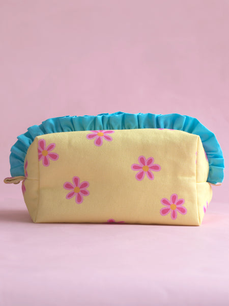 A yellow and pink floral with blue ruffles makeup bag on a pink foreground.