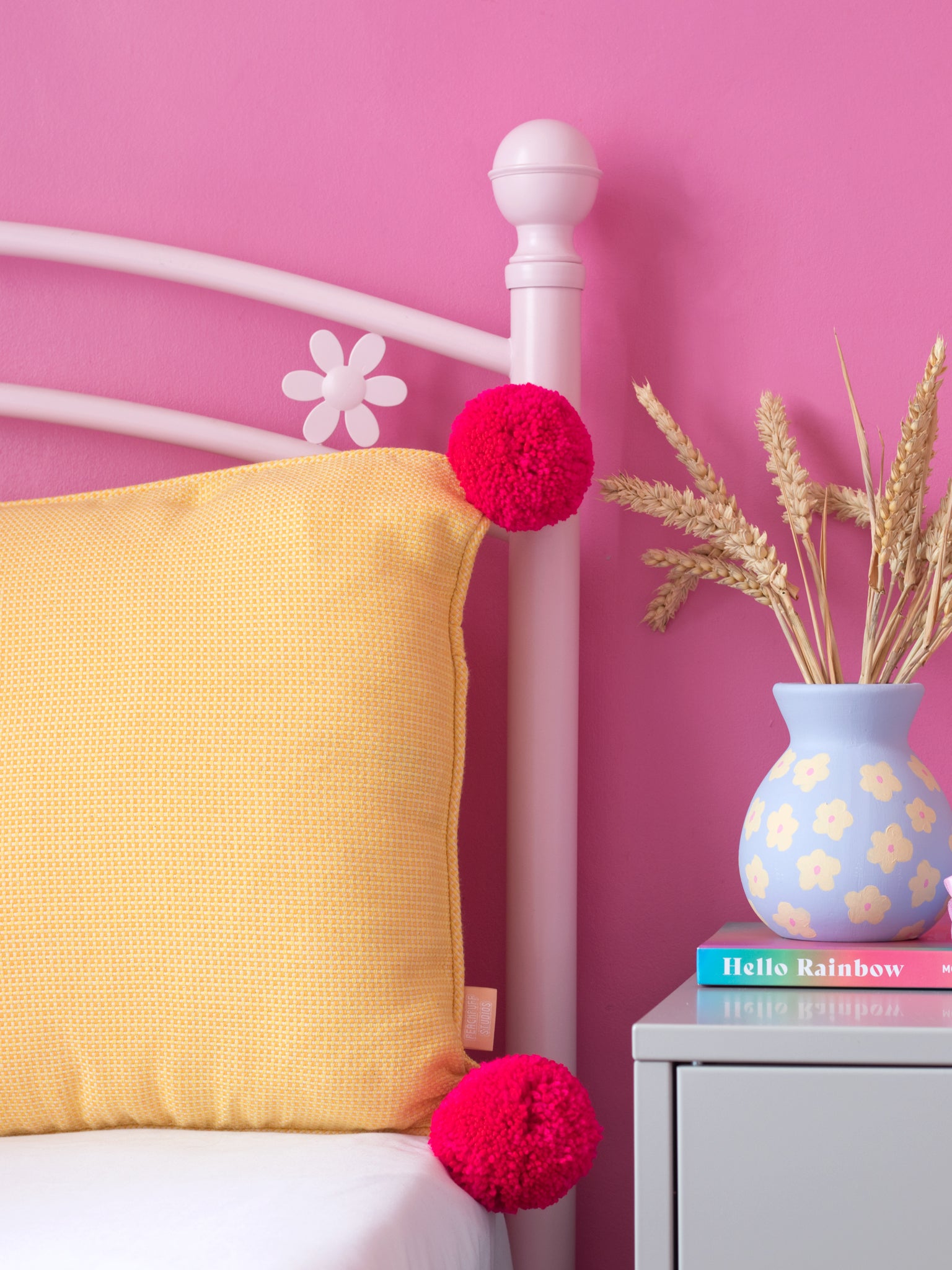 A yellow textured cushion with bright pink pom-poms on a pink flower bed next to a dressing table styled with dried flowers and a book.