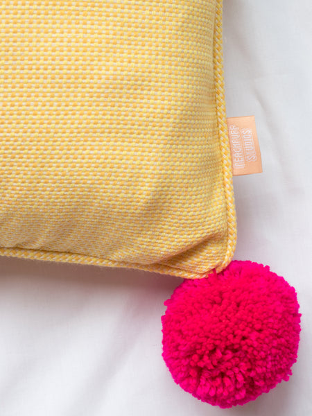 A close-up of a yellow cushion shows the textured fabric and bright pink yarn pom-poms.