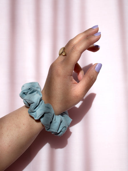 A blue corduroy scrunchie on a female wrist, who is wearing a ring and purple nail varnish.
