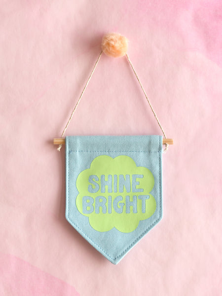 A blue canvas banner that reads Shine Bright in a green flower is hung on a pink wall.