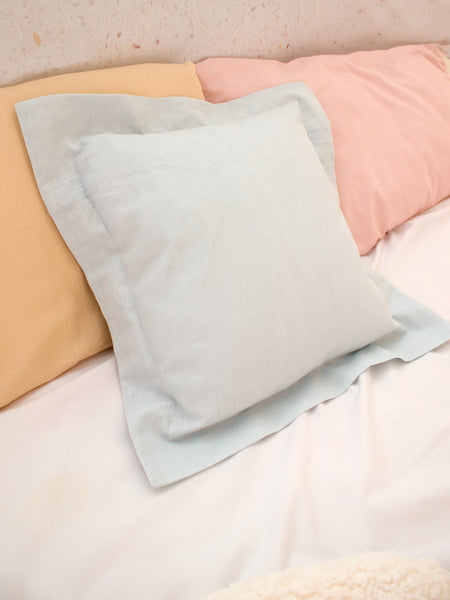 A light blue cotton linen cushion styled on a bed with pastel pillowcases.