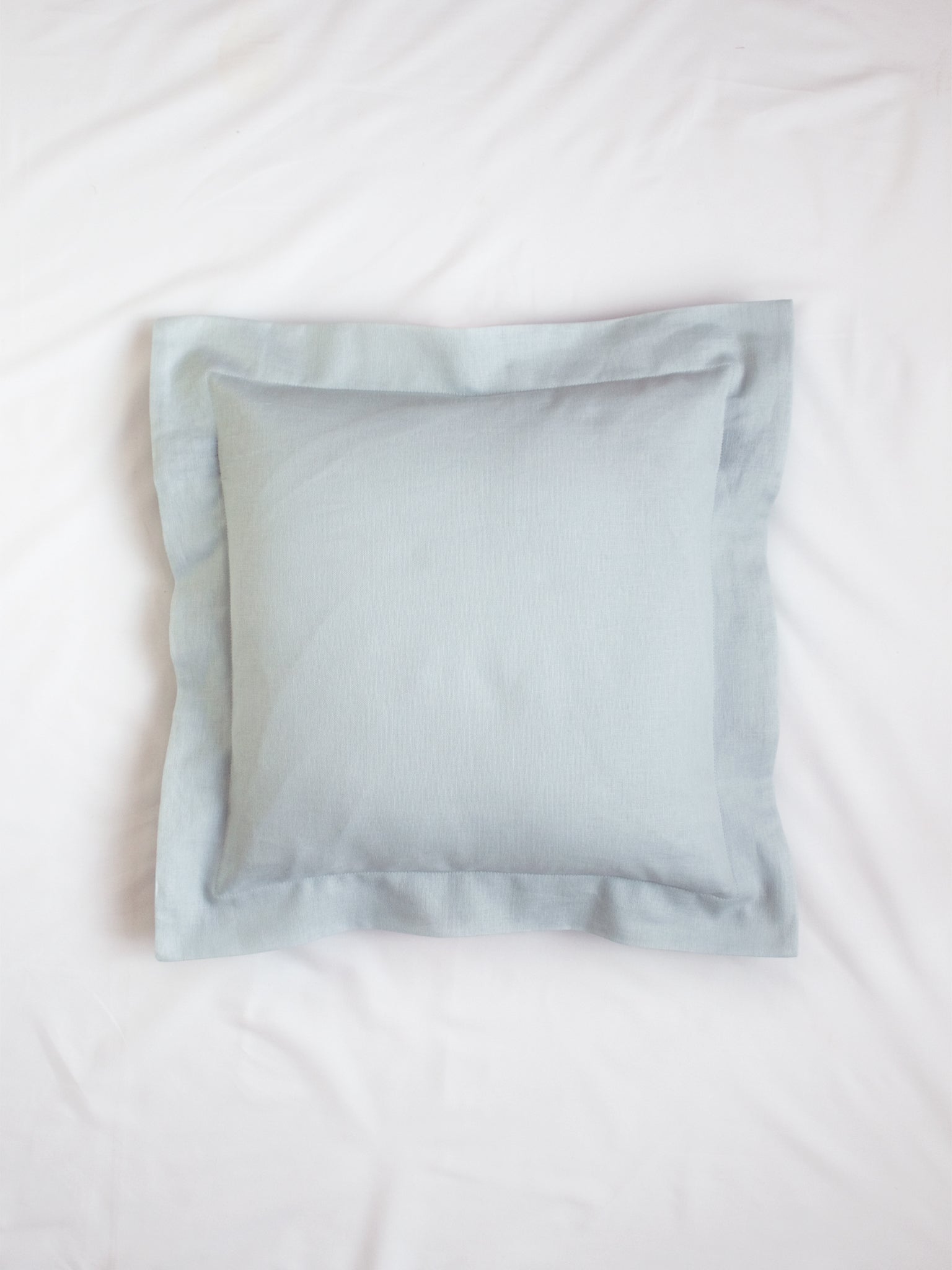 A light blue cotton linen cushion on the centre of a white bedsheet.