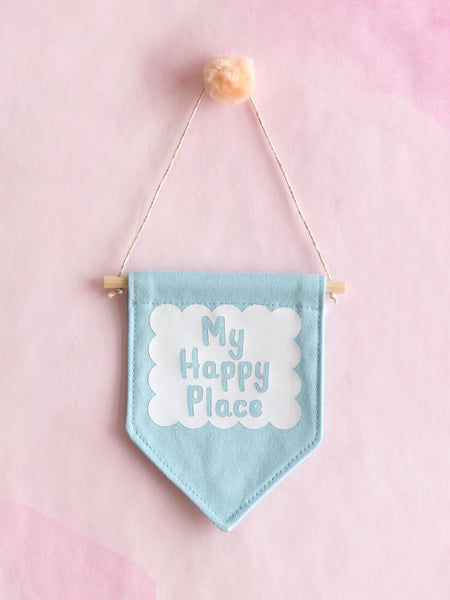 A blue canvas banner that reads My Happy Place in a white scalloped box is hung on a pink wall.