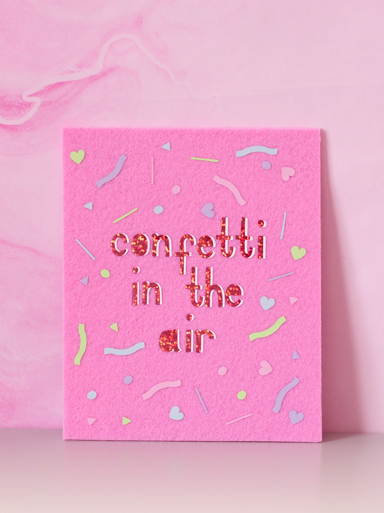 Felt wall art with Confetti In The Air written in glitter vinyl with pastel confetti shapes surrounding the text, sitting in front of a marbled pink wall.