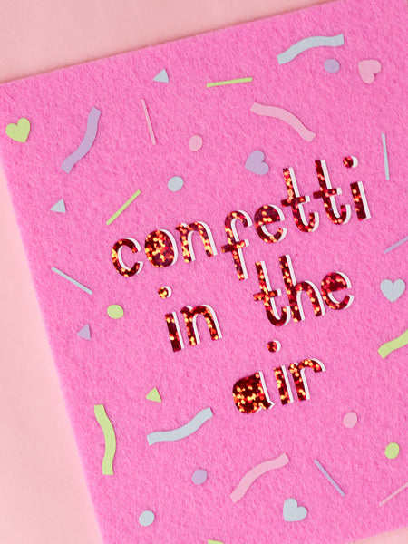 Felt wall art with Confetti In The Air written in glitter vinyl. Pastel confetti shapes surround the text. 