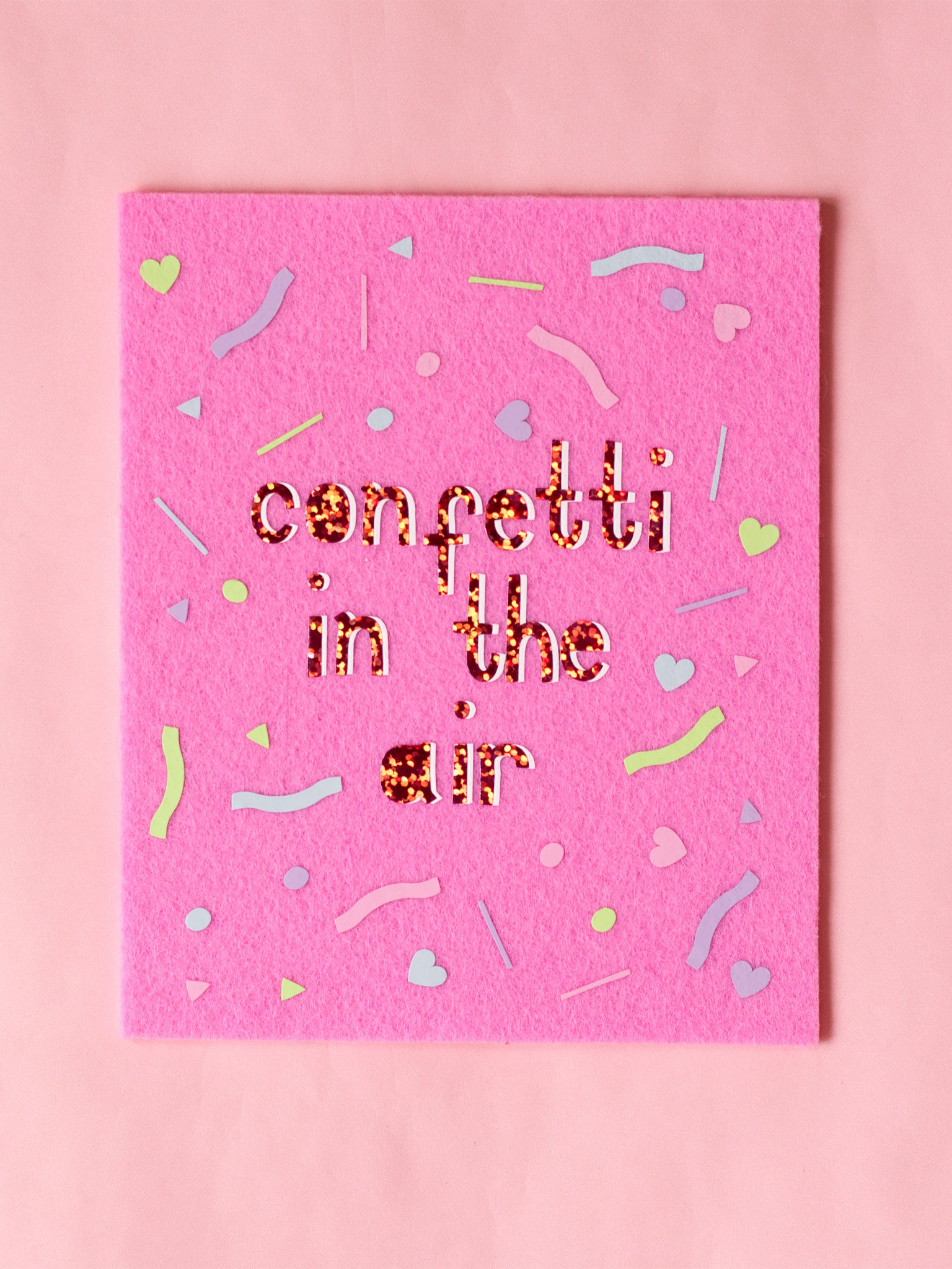 Felt wall art with Confetti In The Air written in glitter vinyl. Pastel confetti shapes surround the text. 
