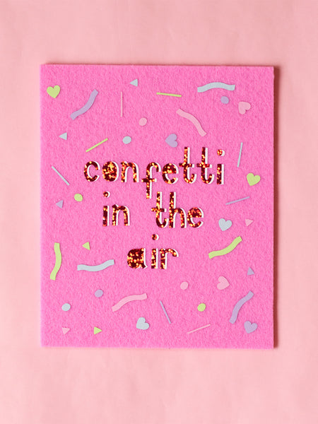 Felt wall art with Confetti In The Air written in glitter vinyl. Pastel confetti shapes surround the text. 