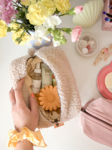 A female hand with a scrunchie on her wrist opens a cream-fluffy makeup bag to reveal beauty products.