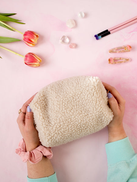 A pair of female hands holding a cream-fluffy zip pouch on a pink-marbled floor. Flowers, crystals, makeup brushes and hair clips offset at the top.