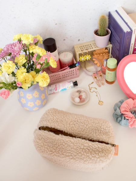 A fluffy cream zip pouch opened on a busy dressing table filled with beauty products, books and a vase of flowers.