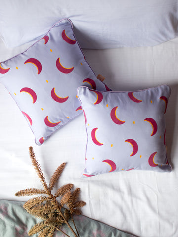 A pair of lilac pink cushions with pink and yellow crescent moons styled on a bed with sheets and flowers.