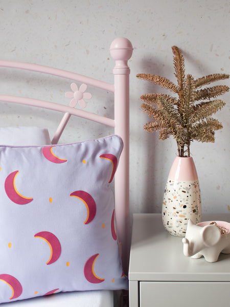 A lilac cushion with pink and yellow vinyl crescent moons on a pink flower bed next to a dressing table styled with flowers and trinkets.