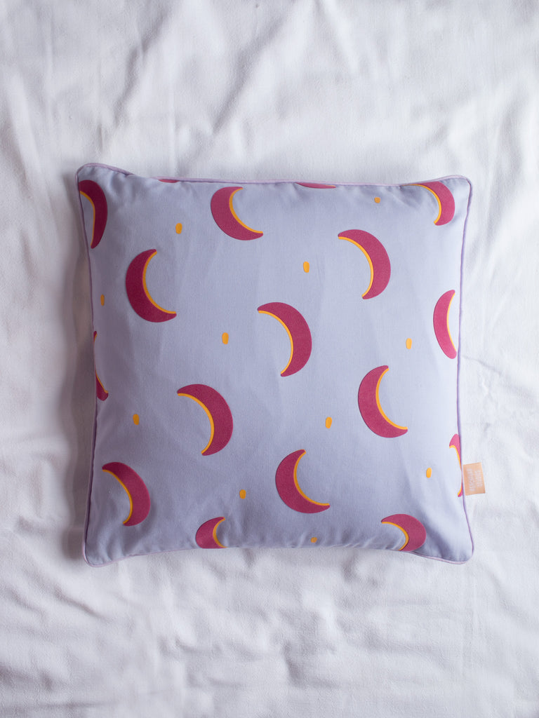 A lilac cushion with pink and yellow vinyl crescent moons on the centre of a white bedsheet.