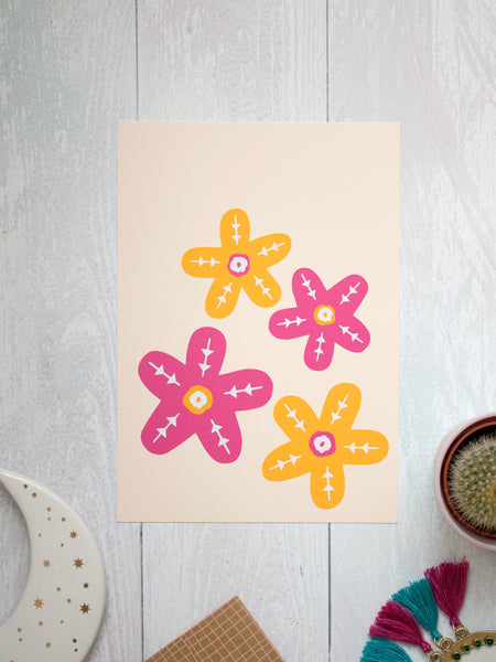 An art print of two orange and two pink flowers lay on a white wooden floor. Trinkets and a cactus lay offset at the bottom.