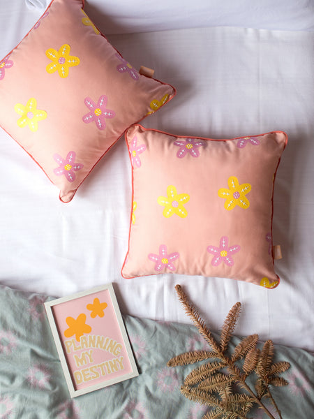 A pair of dusty pink cushions with pink and yellow vinyl flowers styled on a bed with sheets and flowers.