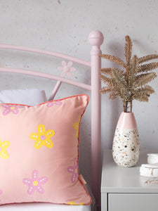 A dusty pink cushion with pink and yellow vinyl flowers on a pink flower bed next to a dressing table styled with flowers and trinkets.