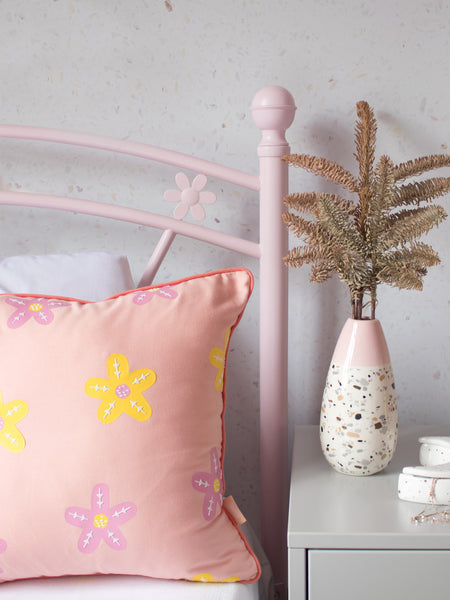 A dusty pink cushion with pink and yellow vinyl flowers on a pink flower bed next to a dressing table styled with flowers and trinkets.