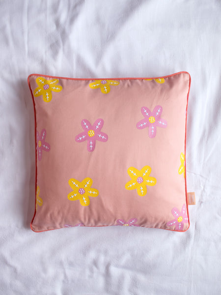 A dusty pink cushion with pink and yellow vinyl flowers on the centre of a white bedsheet.
