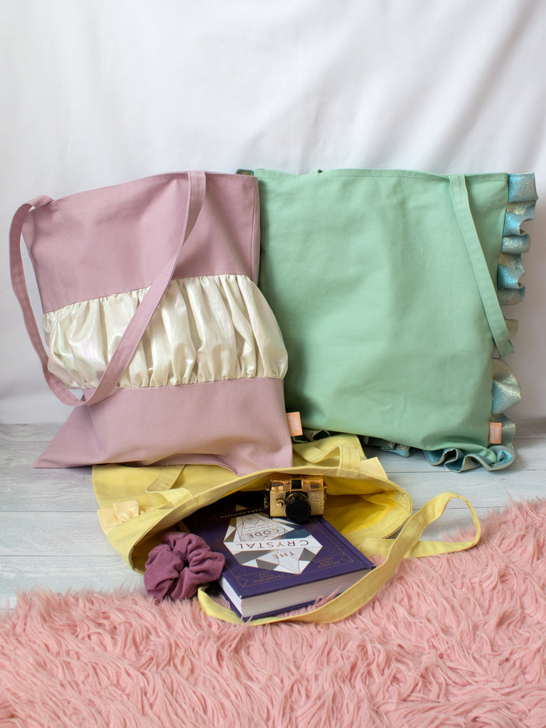 Three different coloured tote bags lay on a white wooden floor. 