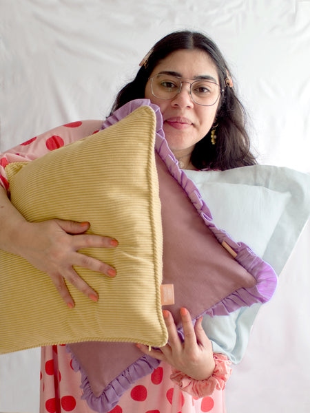 A female holding three different coloured cushions in her arms and simpling in front of a white background.