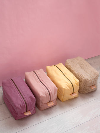 A top view of four different coloured makeup bags lined up on a white wooden floor against a pink wall.