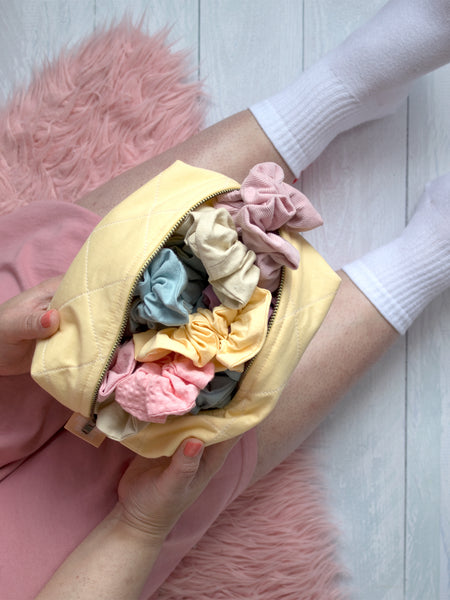 A female is sitting on a pink rug holding a yellow quilted makeup bag in her lap packed with different coloured scrunchies.