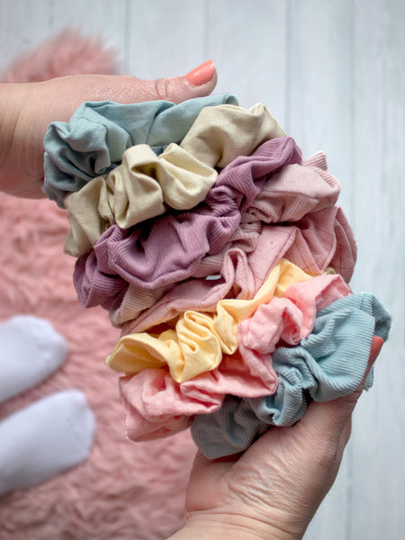 Female hands holding eight different coloured scrunchies above a white wooden floor and a fluffy pink rug.