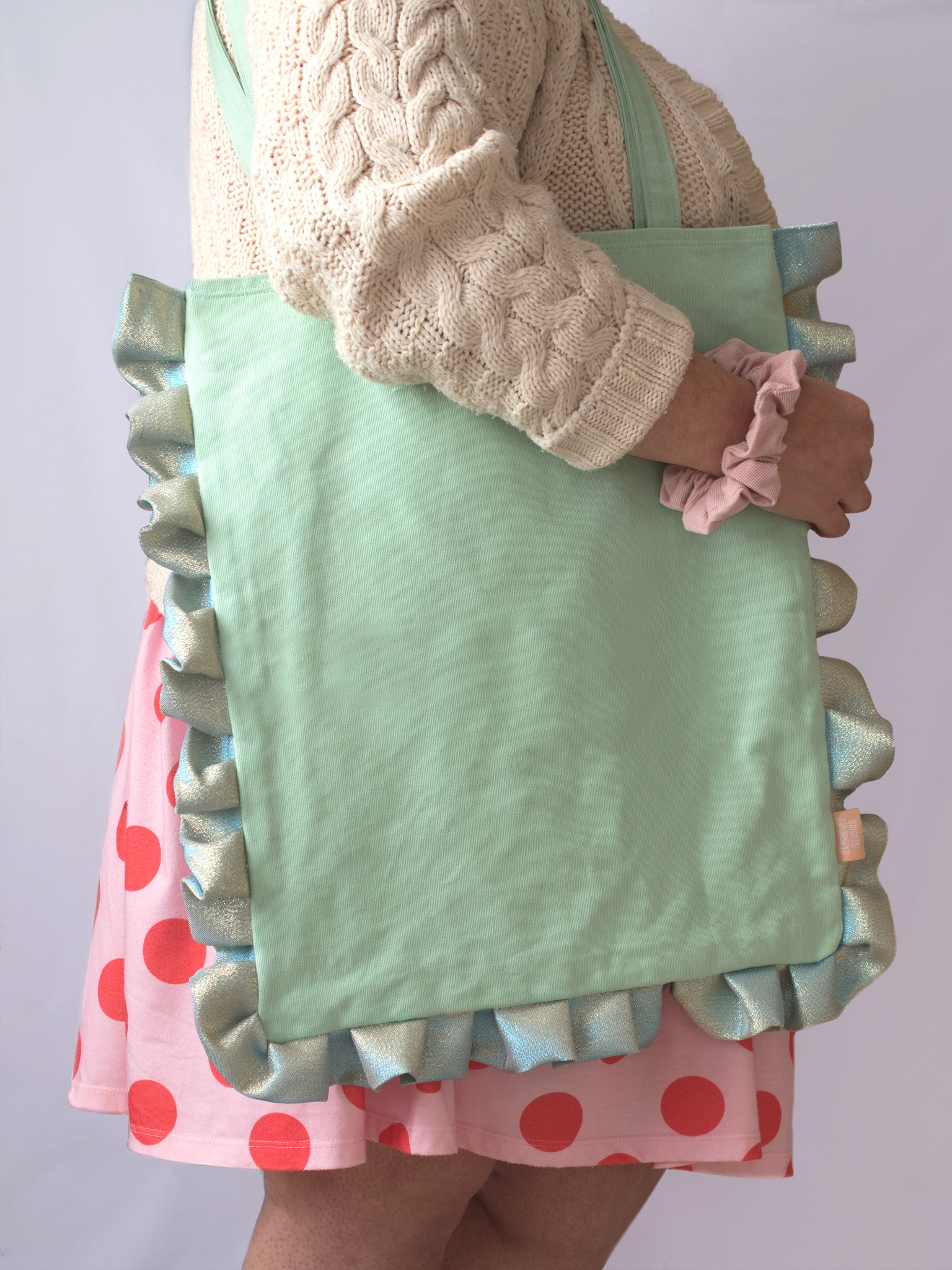 A mint green ruffle tote bag is worn on the shoulder of a female who is wearing a pink and red polka dot dress and cream knit cardigan.