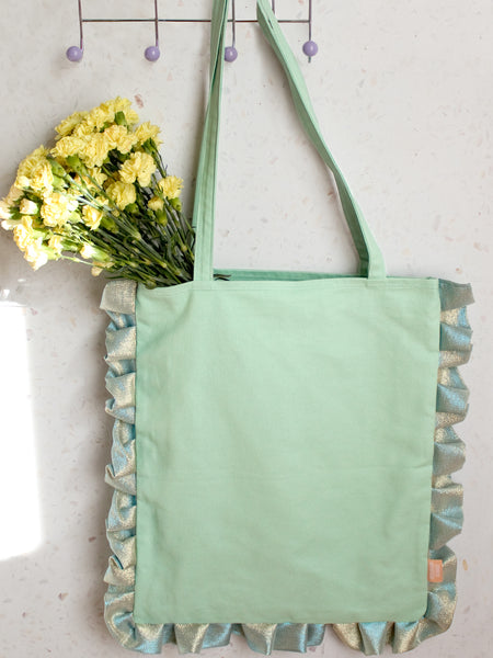 A mint green ruffle tote bag holding yellow flowers is hung on a peg behind a terrazzo wall.