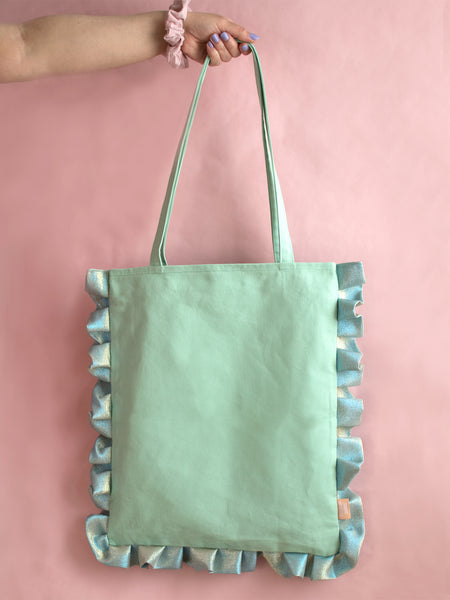 A female arm is holding a mint green ruffle tote bag against a pink foreground.