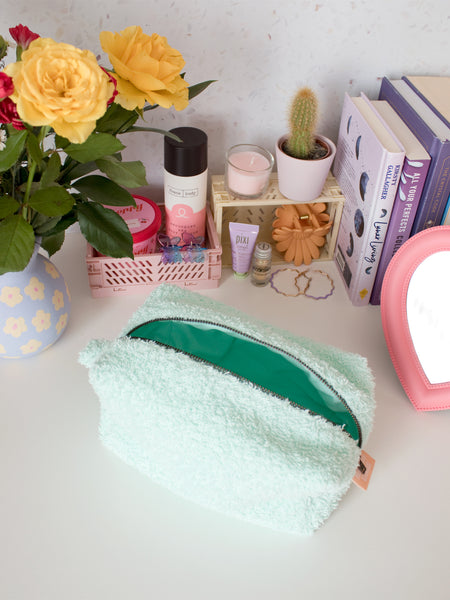 A mint green towelling zip pouch opened on a busy dressing table filled with beauty products, books and a vase of flowers.